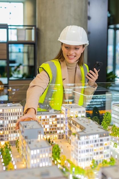 Arquiteto confiante em hardhat usando smartphone enquanto analisa modelo de complexo de escritório no local de trabalho